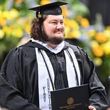 An SNHU graduate in a cap and gown, holding a diploma and wearing a stole that says First-Generation
