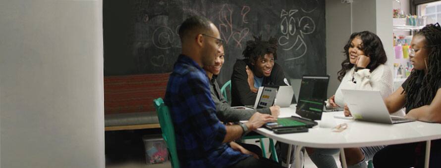 A group of students in a study room working on laptops together and smiling.