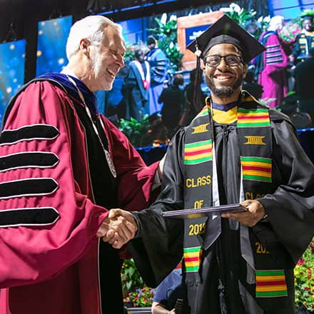 A man in graduation cap and gown shaking hands with a university president as he receives his degree.