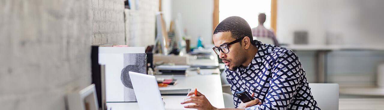A man wearing glasses working on his laptop while holding a smartphone in one hand and a mechanical pencil in the other in an office environment.