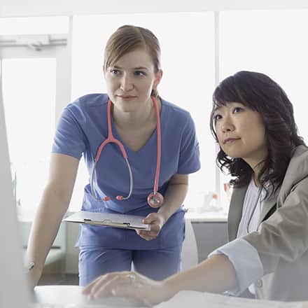 A woman who became a licensed practical nurse, wearing a stethoscope, holding a clipboard and leaning toward a computer screen a healthcare professional is using.