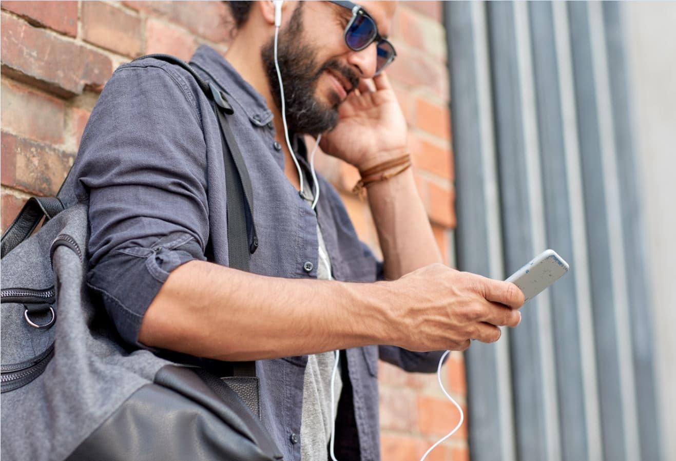 A bearded man wearing sunglasses using headphones to listen to a podcast on his phone.