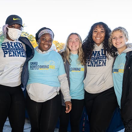 A group of five SNHU students, three wearing SNHU Homecoming shirts and two wearing SNHU Penmen shirts.