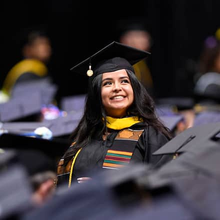 An SNHU graduate in cap and gown, standing among many other sitting graduates.