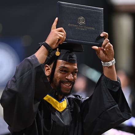 A graduate dressed in a cap and gown and holding a diploma over his head at the Fall 2023 SNHU Commencement.