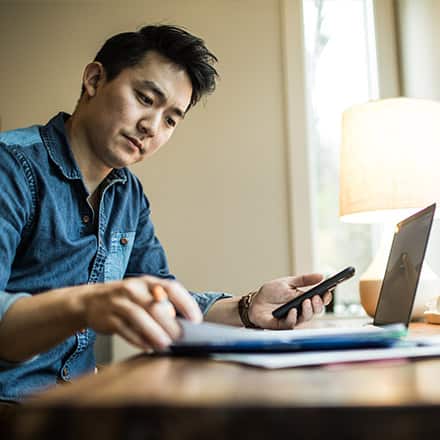 A man holding a cell phone and in front of his laptop as he works remotely.