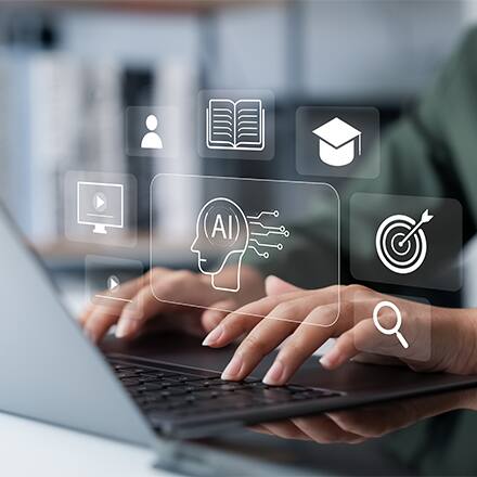 Two hands typing on a laptop with transparent icons depicting artificial intelligence, a book, graduation cap, bullseye target, magnifying glass, play buttons and a person.