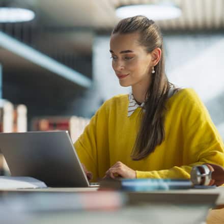 A student wearing a yellow sweater, using a laptop in a public library to earn her master's degree online.