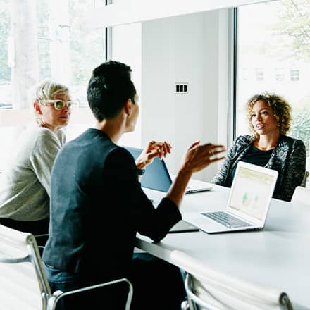 Three coworkers sitting around a table at a meeting with their laptops open for their job they got with their business degree.