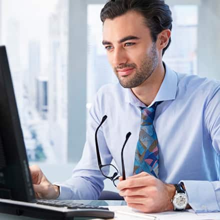 A man with a business degree in a shirt and tie holding his glasses while working at a computer.