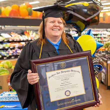 Carol D'Anna in her cap and gown holding her diploma, standing in the center of Walmart produce section
