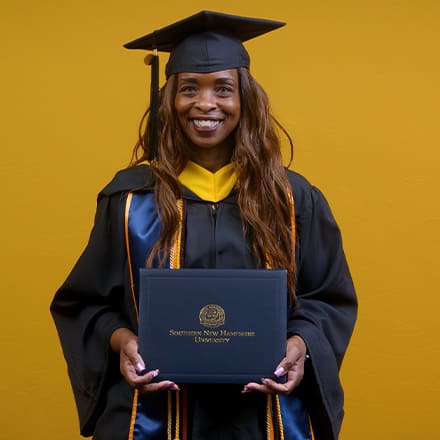 Chantel Hutchison, a 2025 SNHU graduate who earned her bachelor's in public health, holding her diploma and wearing her cap and gown in front of a yellow background.