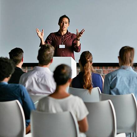 A man with a communications degree at a podium in front of an audience giving an oral presentation.