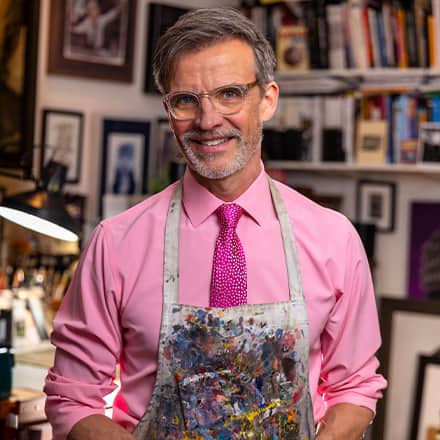 Dale Stephanos, a 2023 graduate from SNHU, standing in his studio and wearing an apron covered in paint.