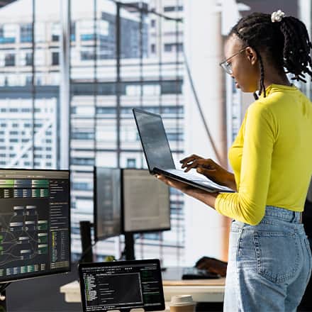 A woman working on a laptop surrounded by other computers and tech equipment to represent database administration.