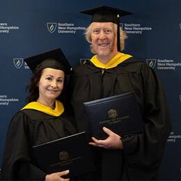 2024 SNHU graduates, David and Irina Roach, who earned their master's in cybersecurity, holding their diplomas, wearing their caps and gowns in front of a blue background.