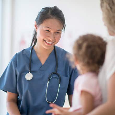 A nursing professional that finished the degree needed to become a nurse, wearing blue scrubs and a stethoscope and meeting with a pediatric patient. 