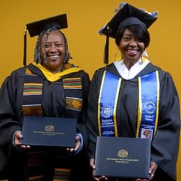 Debra Douglas and Deidra Fransaw holding their diplomas in front of a yellow background at their SNHU commencement ceremony.