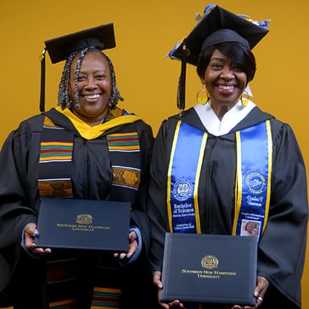 Debra Douglas and Deidra Fransaw holding their diplomas in front of a yellow background at their SNHU commencement ceremony.