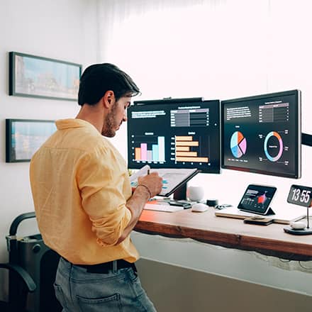 A man in an orange shirt standing at a desk looking at two monitors and researching how to become an economist.