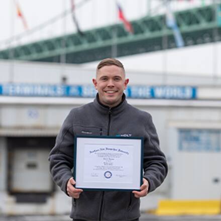 Rob Thomas, a 2024 SNHU graduate holding his diploma standing outside in an industrial area.