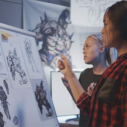 A game designer working with an artist to create a video game character concept using sketches and digital artwork in a design studio.