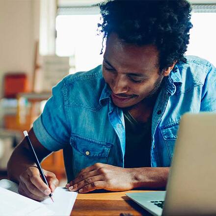 A student wondering how to become a writer, writing in a notebook at a desk with a laptop open in front of him.