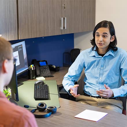 A hiring manager turned away from a computer, speaking to a job candidate about a technical interview.