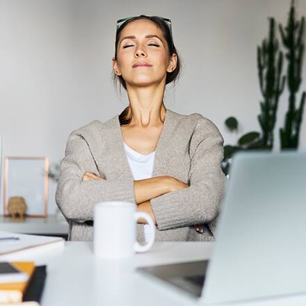 A woman sitting at her desk with her arms crossed and eyes closed practicing mindfulness meditation.