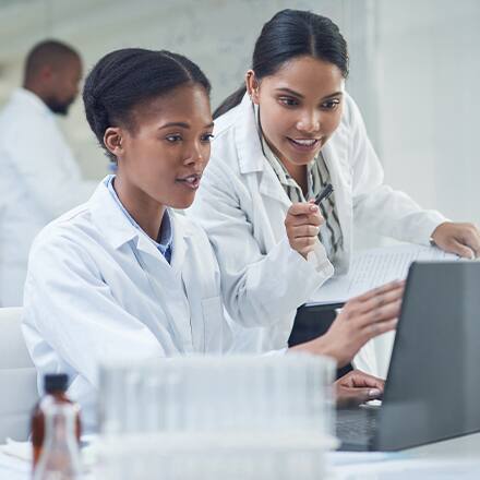 Two women in lab coats working in health science looking at a computer with a co-worker in the lab behind them