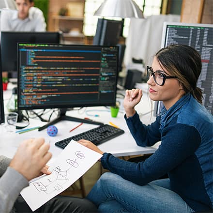 A computer science professional discussing a workflow diagram with a colleague who has code on her desktop.