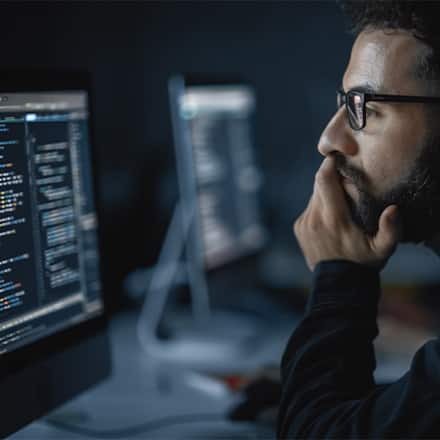 A man wearing glasses looks at code on a computer screen with his chin resting on his hand.