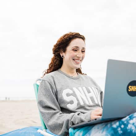 Mariel Embry, a 2022 SNHU graduate sitting on a beach with her laptop working on her liberal arts degree.