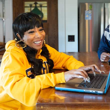 Remy Twine, a 2025 SNHU marketing graduate wearing a yellow SNHU sweatshirt working on a laptop at a wooden table inside a home, with another person seated nearby.