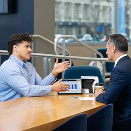 Two people seated at a table during a business meeting, having a discussion, with a tablet displaying charts on the table between them.