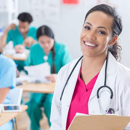 A nurse educator wearing a white lab coat and stehoscope in a classroom with nursing students behind her.