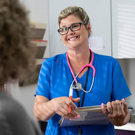 An ethical nurse wearing blue scrubs and a pink stethoscope speaking to a patient.