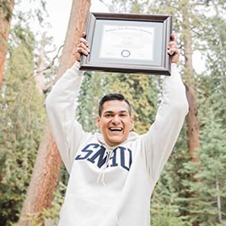 Juan Munoz, a 2022 and 2024 SNHU graduate, holding the diploma for his MBA over his head with pine trees behind him.