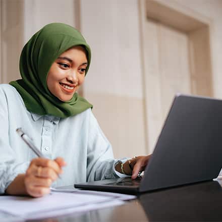 A smiling woman wearing a green hijab and a button-down shirt sitting at a laptop with a pen in hand.