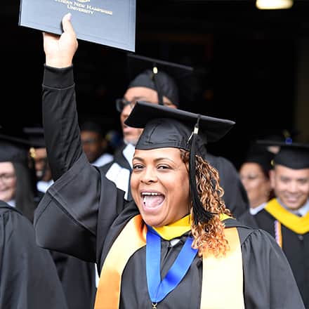 An SNHU Class of 2023 graduate dressed in a cap and gown and holding a diploma cover up in the air.