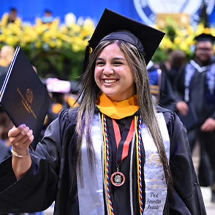 A smiling graduate at SNHU’s Spring 2026 commencement ceremony while holding their diploma, wearing a cap and gown with honors cords.