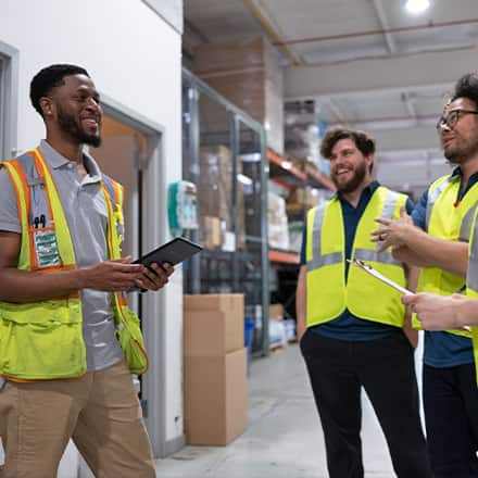 A supply chain manager holding a tablet, speaking with other professionals wearing fluorescent yellow vests and working in a warehouse.