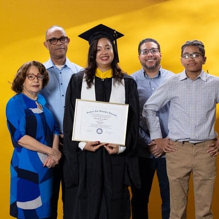 Brendaliz De Los Santos, center, dressed in a graduation cap and gown and holding her SNHU diploma, surrounded by 4 family members who supported her in college.