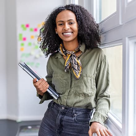 A woman smiling while holding a laptop and notebooks in a modern office.