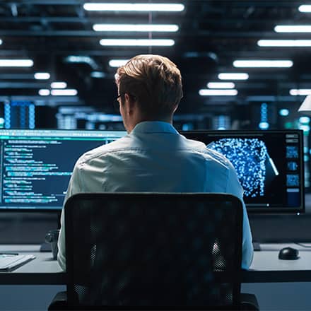 The back of a professional with a master's in cybersecurity at work, sitting at a desk in a large office building with two monitors in front of him.