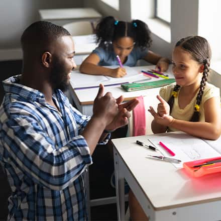 A male teacher kneeling and making eye contact while instructing a child in a classroom.