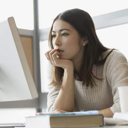 A college student searching for different types of scholarships on a desktop computer with a coffee cup and notebook open beside her.