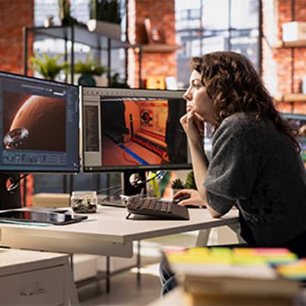 A game designer sitting in front of two monitors with a video game development software open, showing a planet and the inside of a spacecraft.