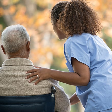 A CNA wearing blue scrubs with her hand on the shoulder of a patient in a wheelchair.