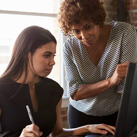 One woman sitting look at a desktop with another woman standing over her looking at the desktop to